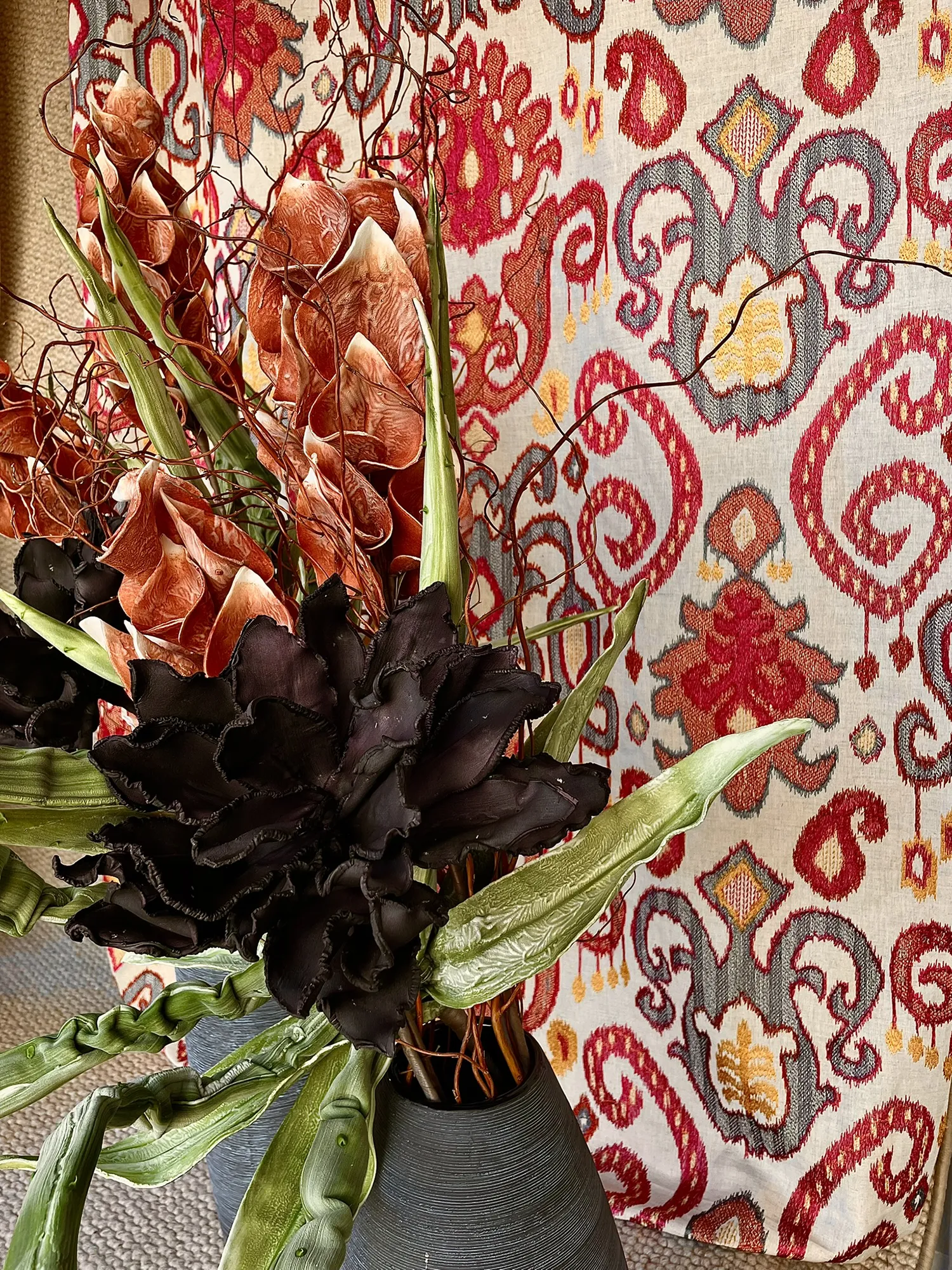 Ein Strauß aus dunklen und rostfarbenen Blumen mit grünen Blättern in einer strukturierten Vase. Der Hintergrund ist mit einem roten und gelben, verschnörkelten Textilmuster versehen. Lebendig und eklektisch.
