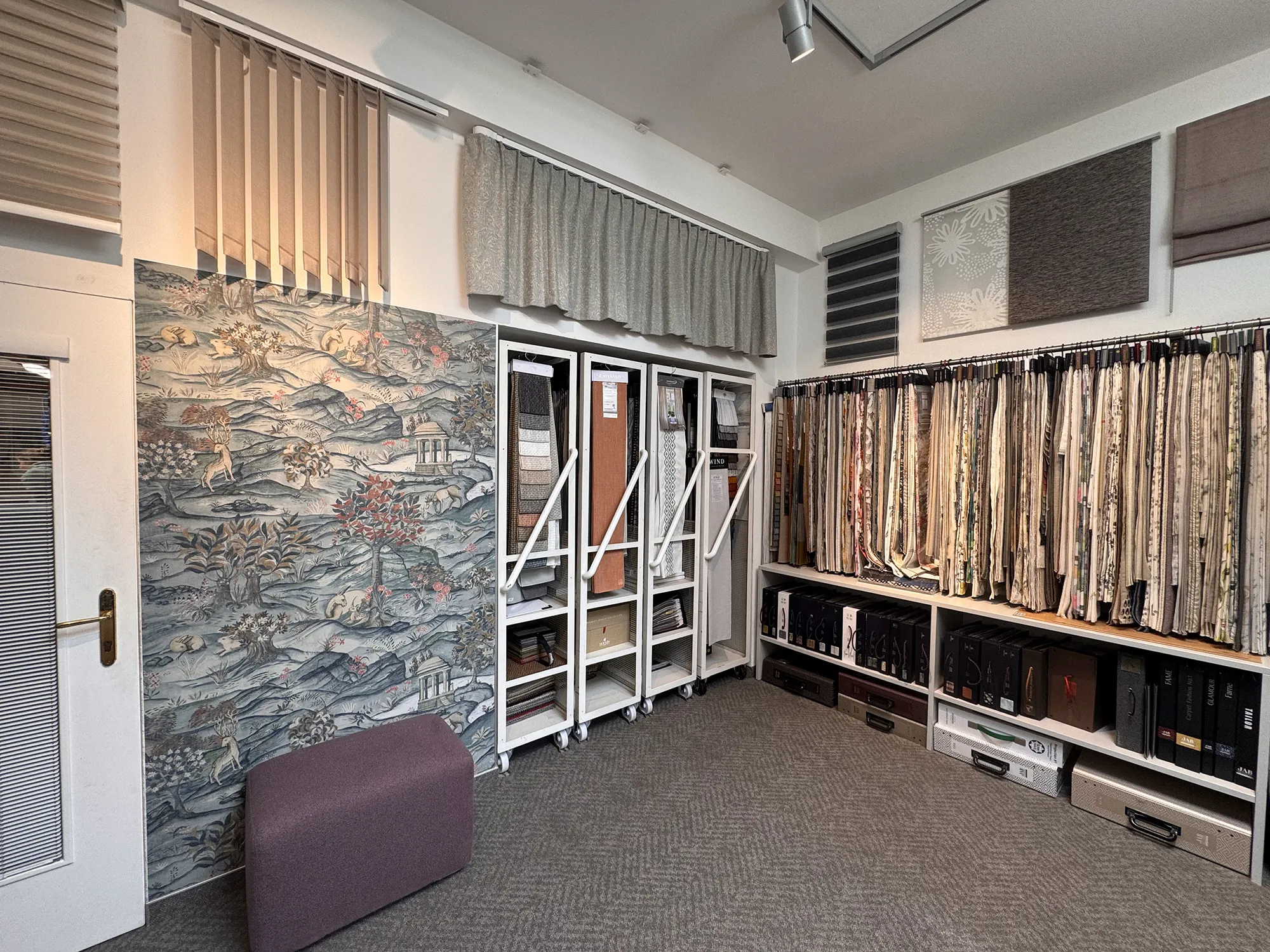 Stoffgeschäft mit hängenden Textilien von Wand zu Wand, bunt gemustertem Stoff auf der linken Seite, Vorhangmustern und geordneten Regalen darunter. Gemütliches Ambiente.