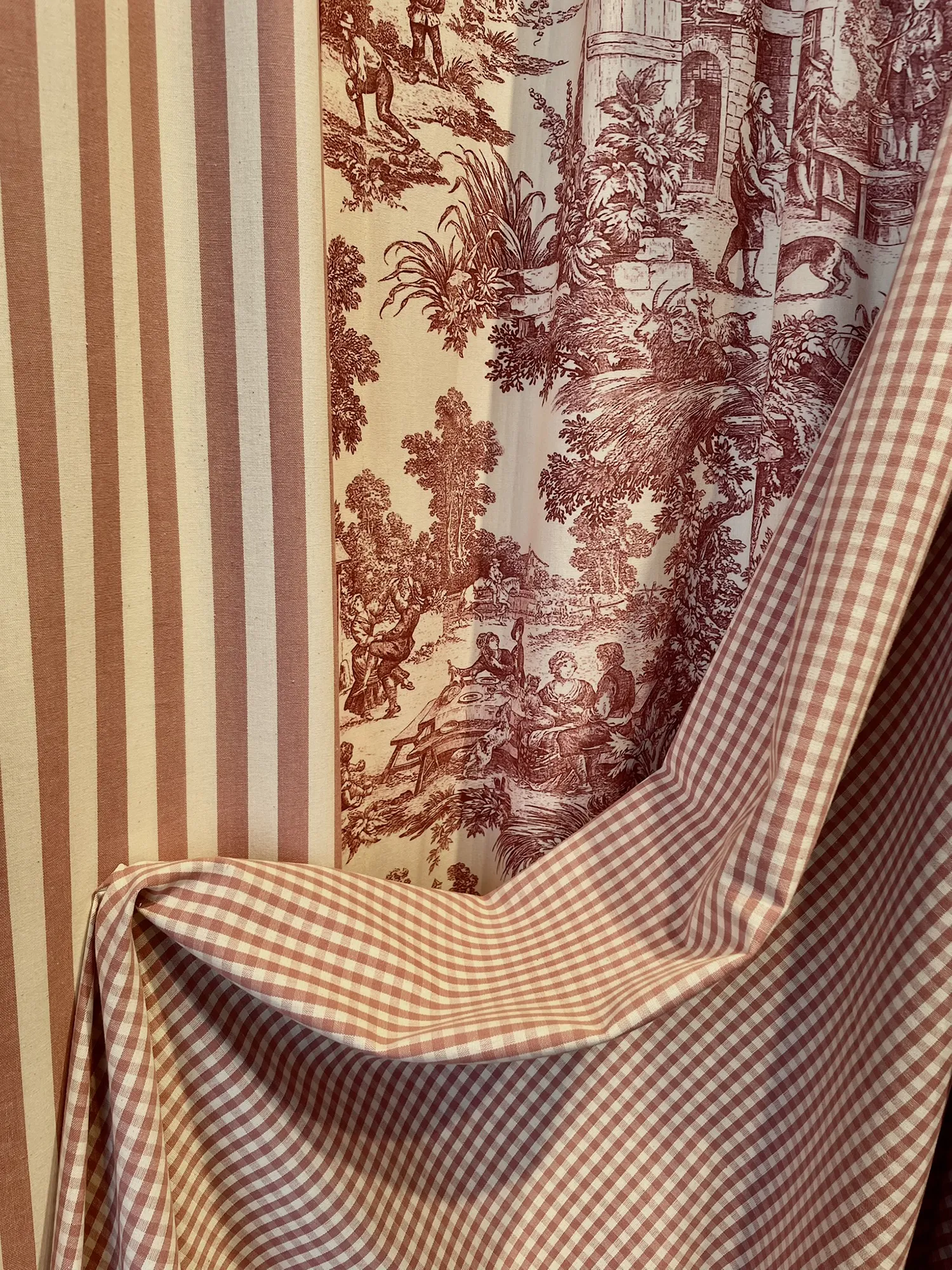 Textilmuster mit roten und weißen Streifen, karierten Gingham-Mustern und einem Toile-Design mit pastoralen Szenen. Die Stoffe vermitteln ein gemütliches Vintage-Gefühl.