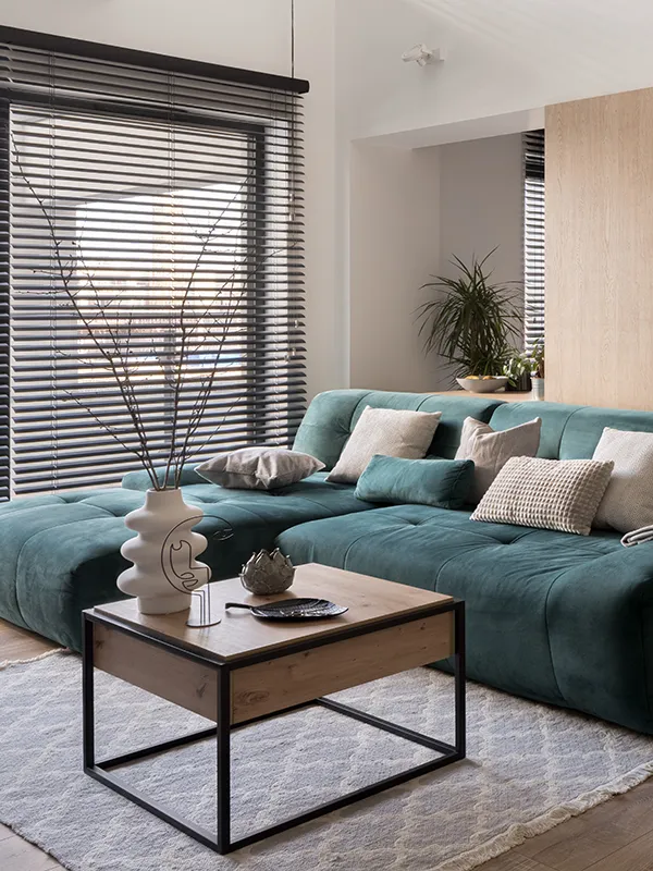 Stilvolles Wohnzimmer mit einem plüschigen tealfarbenen Sektionssofa mit neutralen Kissen. Auf einem modernen Couchtisch aus Holz stehen dekorative Vasen. Große Fenster mit Jalousien lassen natürliches Licht herein und schaffen ein gemütliches Ambiente.