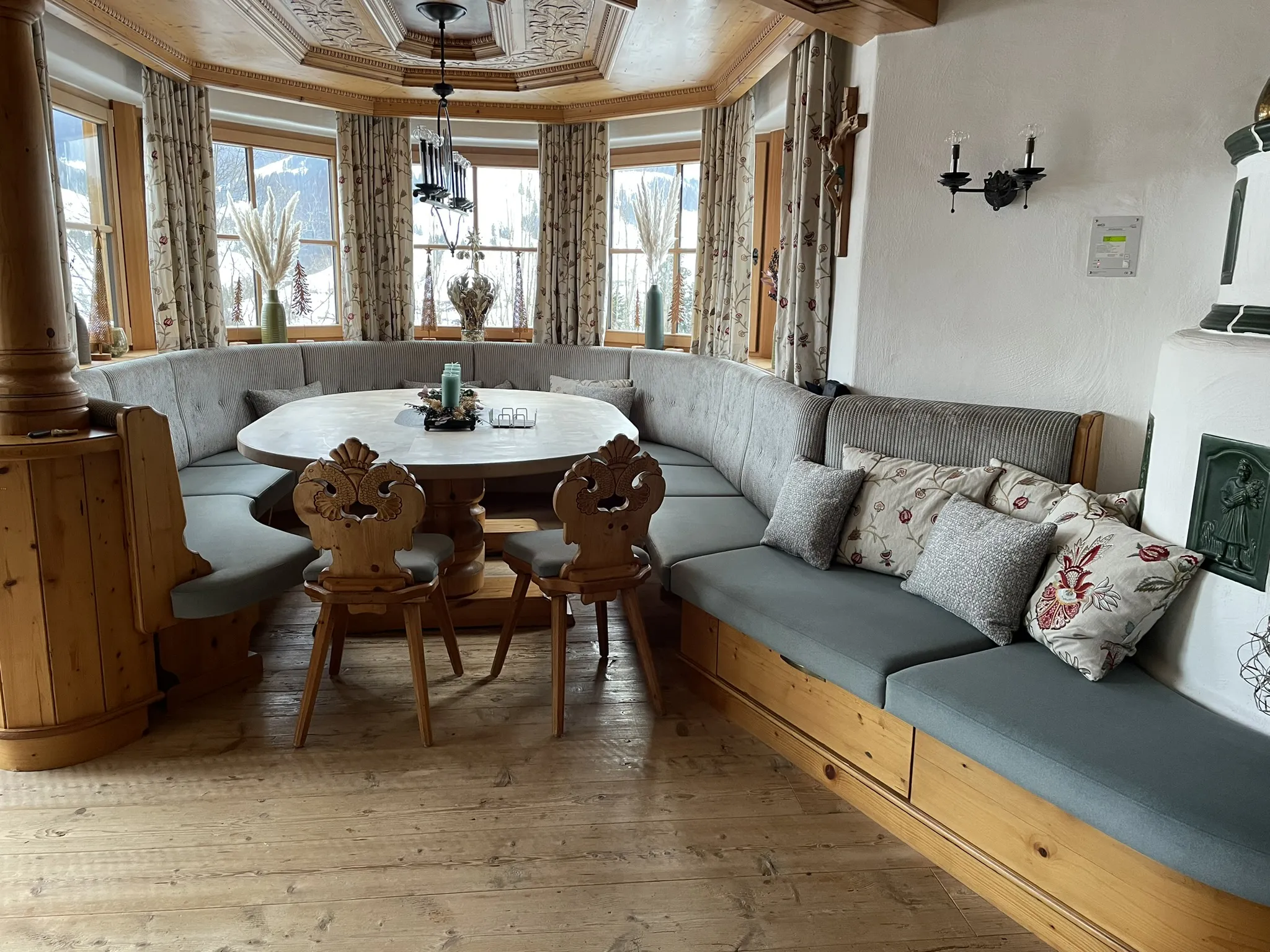 Gemütliche Essecke aus Holz mit rundem Tisch und grauen Sitzpolstern, umgeben von Fenstern mit geblümten Vorhängen. Dekorative Kissen sorgen für Gemütlichkeit.