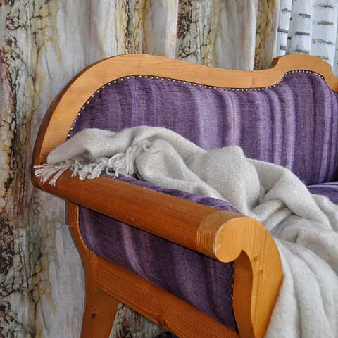 Holzbank mit lila Polsterung und Messingnägeln, drapiert mit einer cremefarbenen Fransendecke. Im Hintergrund befinden sich erdig gemusterte Vorhänge. Gemütliches Ambiente.