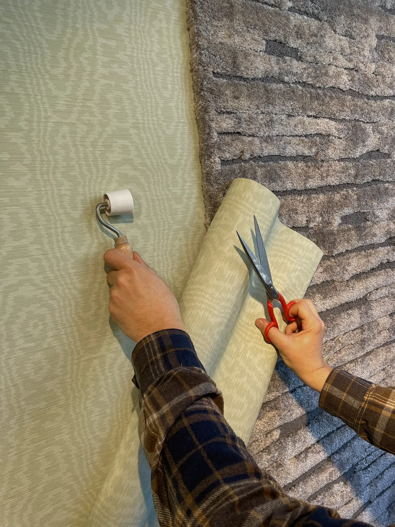 Hände in einem karierten Hemd glätten hellgrüne Tapeten mit einer Rolle und schneiden sie mit einer Schere zu, was einen Kontrast zu einer strukturierten grauen Wand bildet.